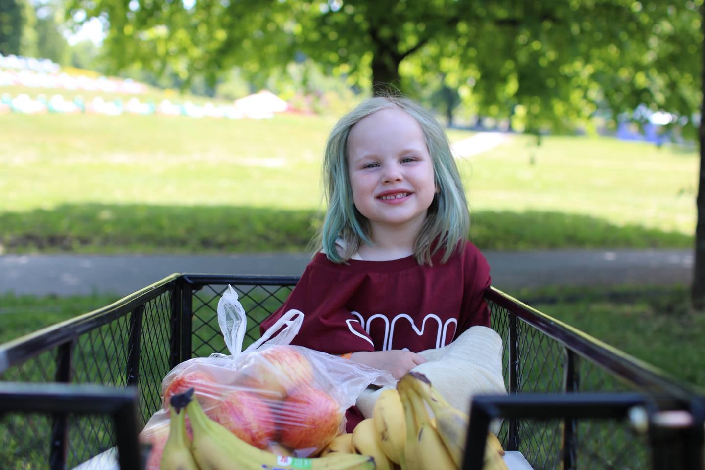 En jente på 4 år som sitter i en vogn med frukt. Hun har blått hår.2