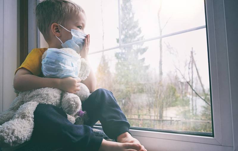Illness child on home quarantine. Boy and his teddy bear both in protective medical masks sits on windowsill and looks out window. Virus protection, coronavirus pandemic, prevention epidemic.