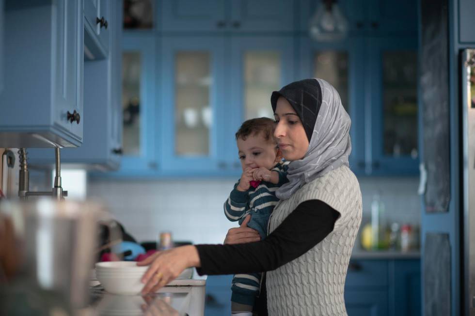 A young Muslim mother holds her infant son on her hip as she works away in the kitchen clearing dishes.  She is dressed casually and wearing a Hijab