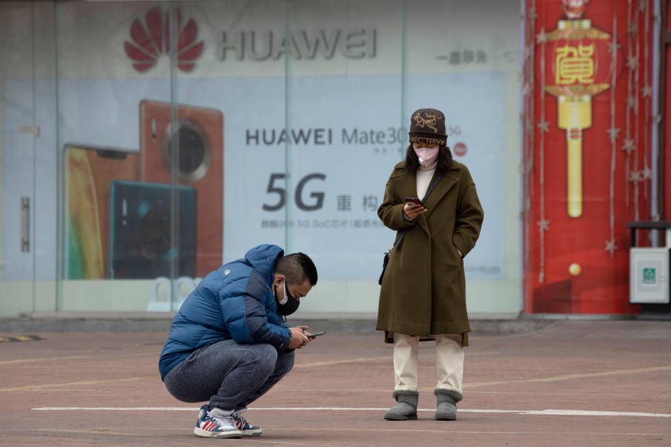 In this Sunday, March 8, 2020, photo, residents wearing masks look at their smartphones near an advertisement for 5G smartphones from Chinese tech giant Huawei in Beijing. Factories in China, struggling to reopen after the coronavirus shut down the economy, face a new threat from U.S. anti-disease controls that might disrupt the flow of microchips and other components they need. For most people, the new coronavirus causes only mild or moderate symptoms. For some it can cause more severe illness. (AP Photo/Ng Han Guan)