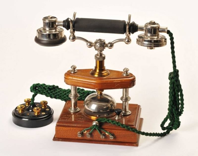 Telefonapparat fra 1902. Åpent bordapparat med elektromagnetisk mikrofon, høretelefon, snor og rosett. De første telefonene var vakkert håndverk av tre og ornamentert metall. Fra Elektrisk Bureau, som hadde produsert telefonapparater sida 1885.