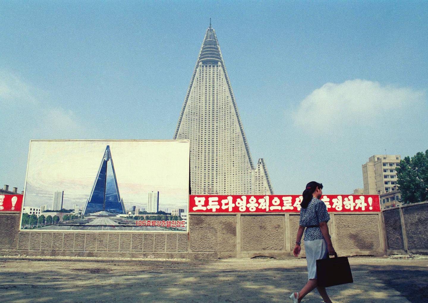 FILE - In this Aug. 22, 1990, file photo, a woman walks past the site of the Ryugyong Hotel under construction in Pyongyang, North Korea. Sign in red reads "Let's all together struggle heroically!" Walls set up to keep people out of a construction area around the gargantuan Ryugyong Hotel were pulled down in July 2017 as the North marked the anniversary of the Korean War armistice to reveal two broad new walkways leading to the building and the big red propaganda sign declaring that North Korea is a leading rocket power. (AP Photo/Vincent Yu, File)