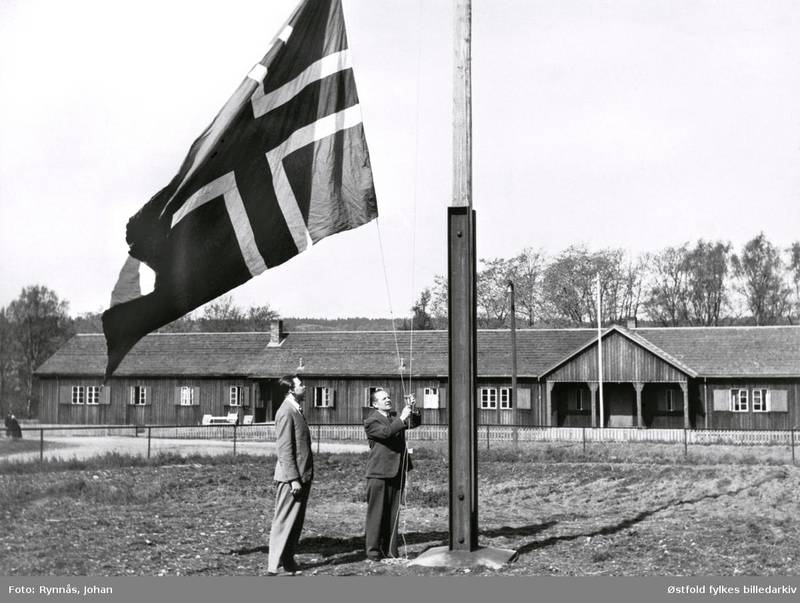 Rådmann Alf Herland og Arne Kinander heiser flagg på frigjøringsdagen 8. mai 1945. I bakgrunnen en Todtbrakke.