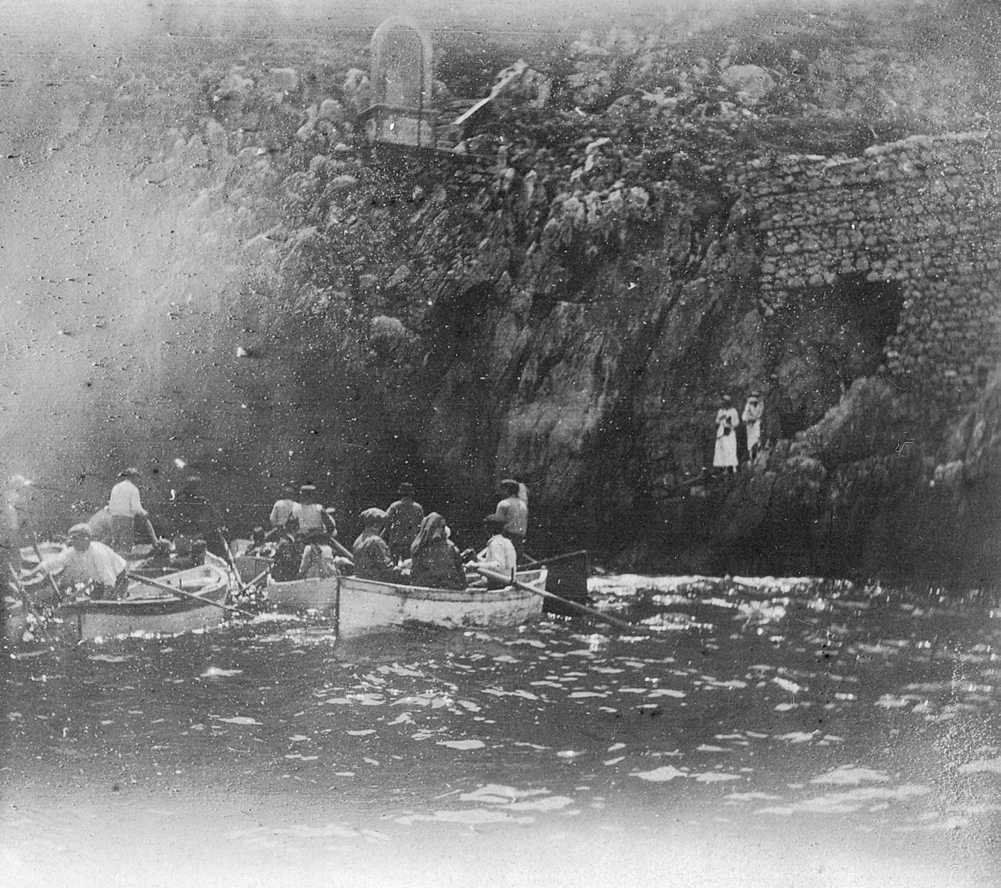 Fotografia in bianco e nero di persone su diverse barche a remi dirette verso una grotta in riva al mare.