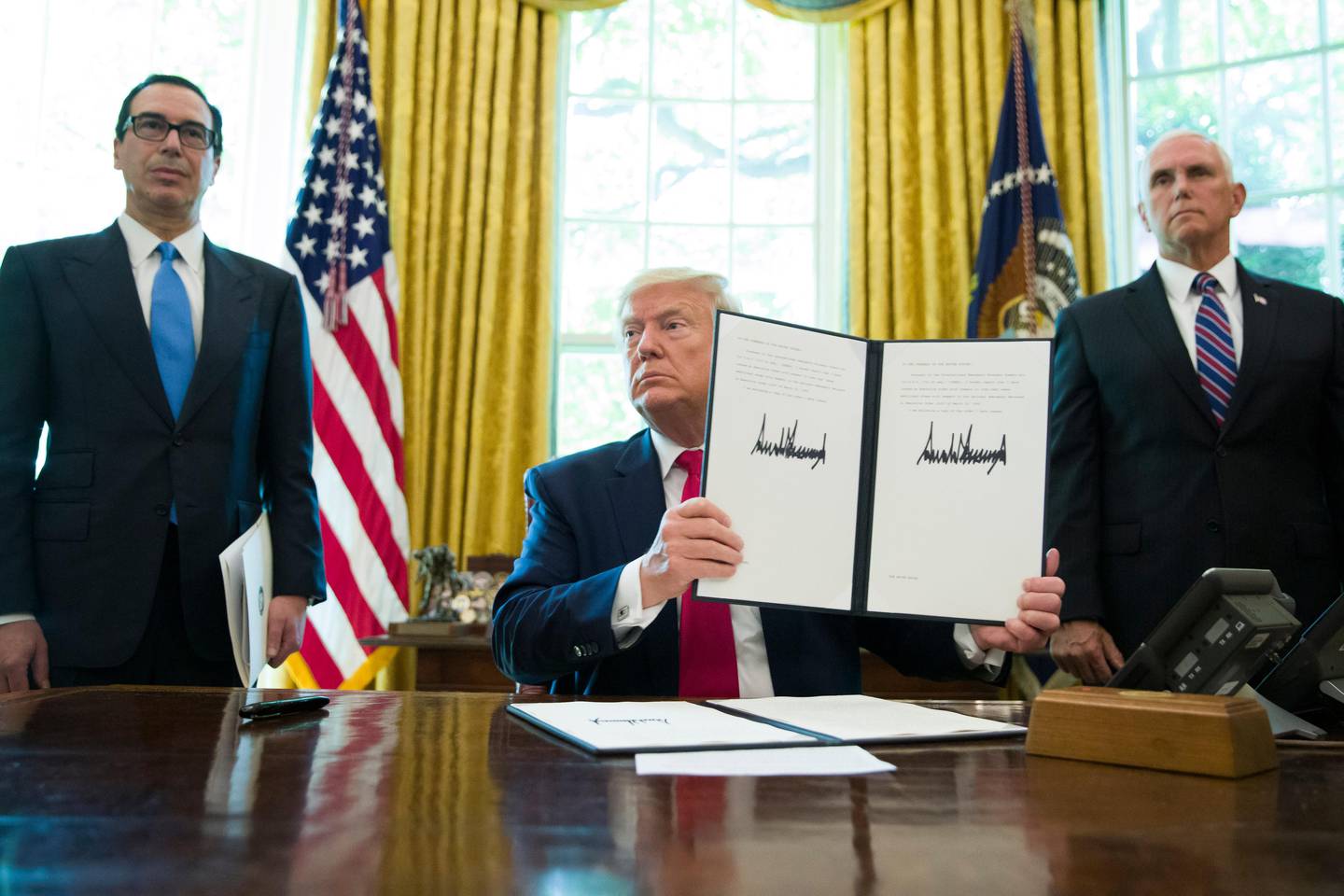 FILE - In this Monday, June 24, 2019 file photo, President Donald Trump holds up a signed executive order to increase sanctions on Iran, accompanied by Treasury Secretary Steve Mnuchin, left, and Vice President Mike Pence, in the Oval Office of the White House, in Washington. A year after President TrumpÄôs unilateral withdrawal from the 2015 deal, the U.S. and Iran are already locked in a volatile standoff. (AP Photo/Alex Brandon, File)