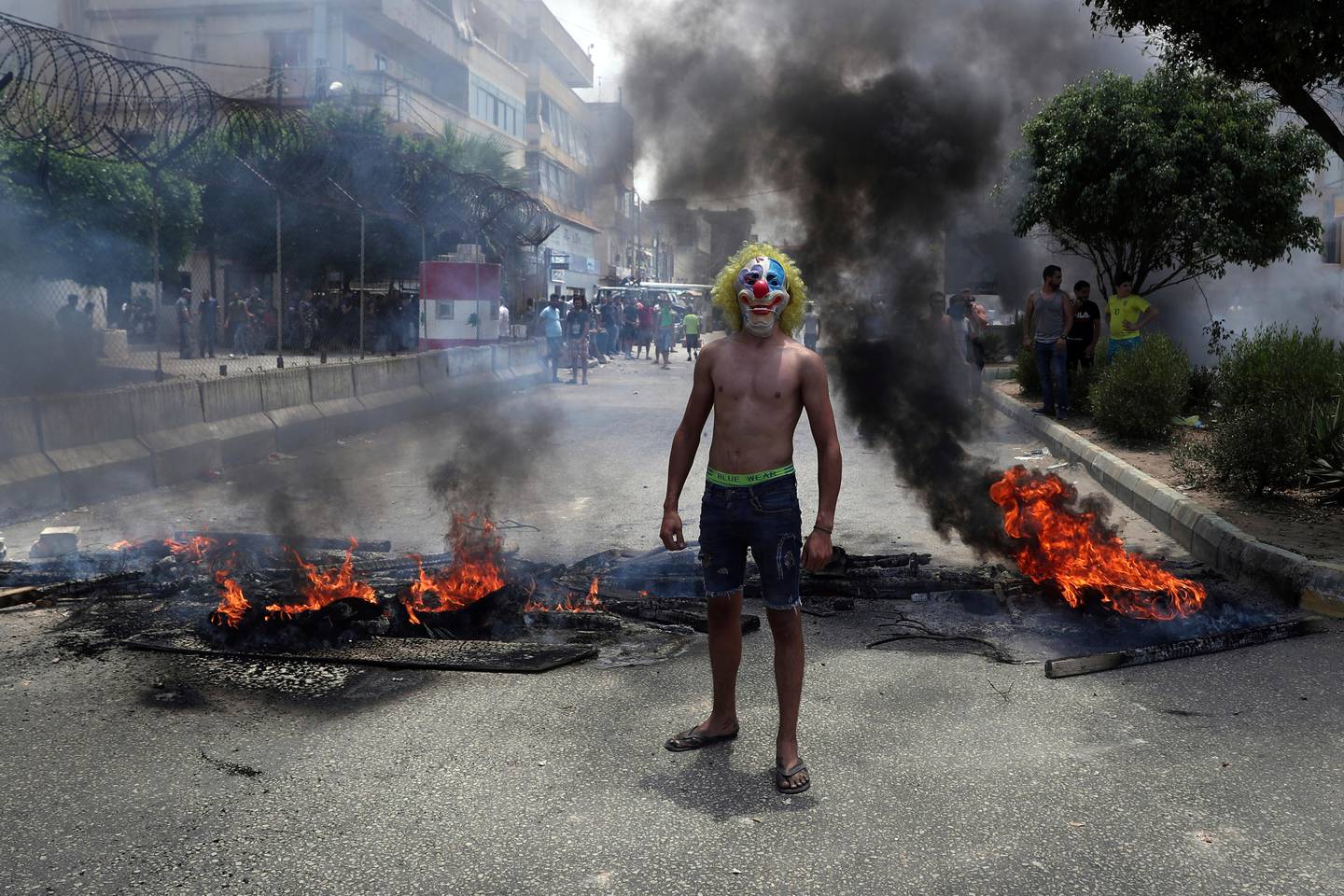 An anti-government demonstrator poses with a clown mask as others burn tires and wood to block a road in Beirut, Lebanon, Tuesday, July 14, 2020. Trash has been piling up in different parts of Lebanon recently as no deal has been reached between the government and waste management companies over payments for their employees. Most workers at the companies are foreigners and want to get paid in U.S. dollars. Lebanon is witnessing shortage in hard currency and the Lebanese pound has lost more than 80% of its value in recent months. (AP Photo/Bilal Hussein)