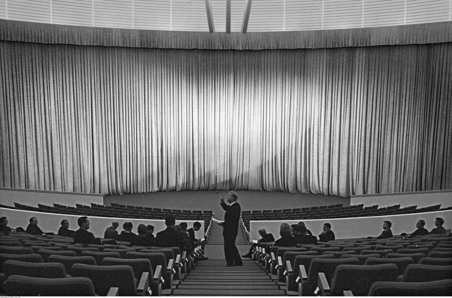 Et sceneteppet er trukket foran scenen. I salen står en mann i midtgangen og peker. I salen sitter det flere menn på de første radene. De ser på mannen som peker.