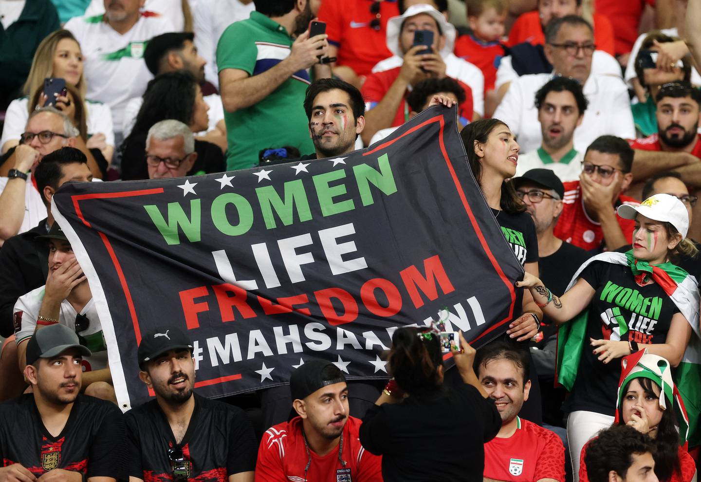 Iran-fans holder et banner med teksten «kvinner, liv, frihet» og #MahsaAmini under fotball-VM i Qatar.