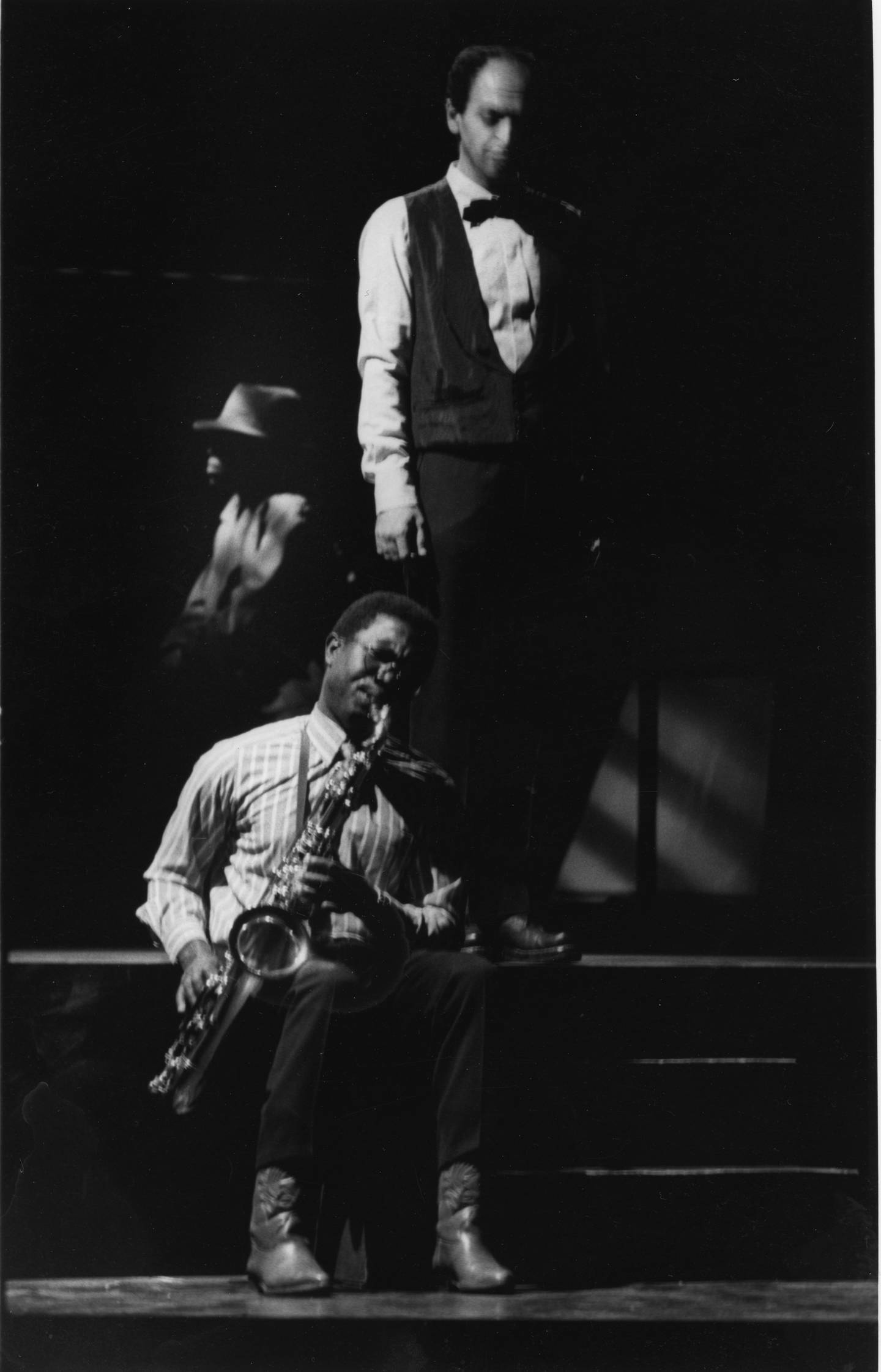 En mann sitter og spiller saksofon. Bak ham står en mann og ser ned. I bakgrunnen synes en mann med hatt i profil.