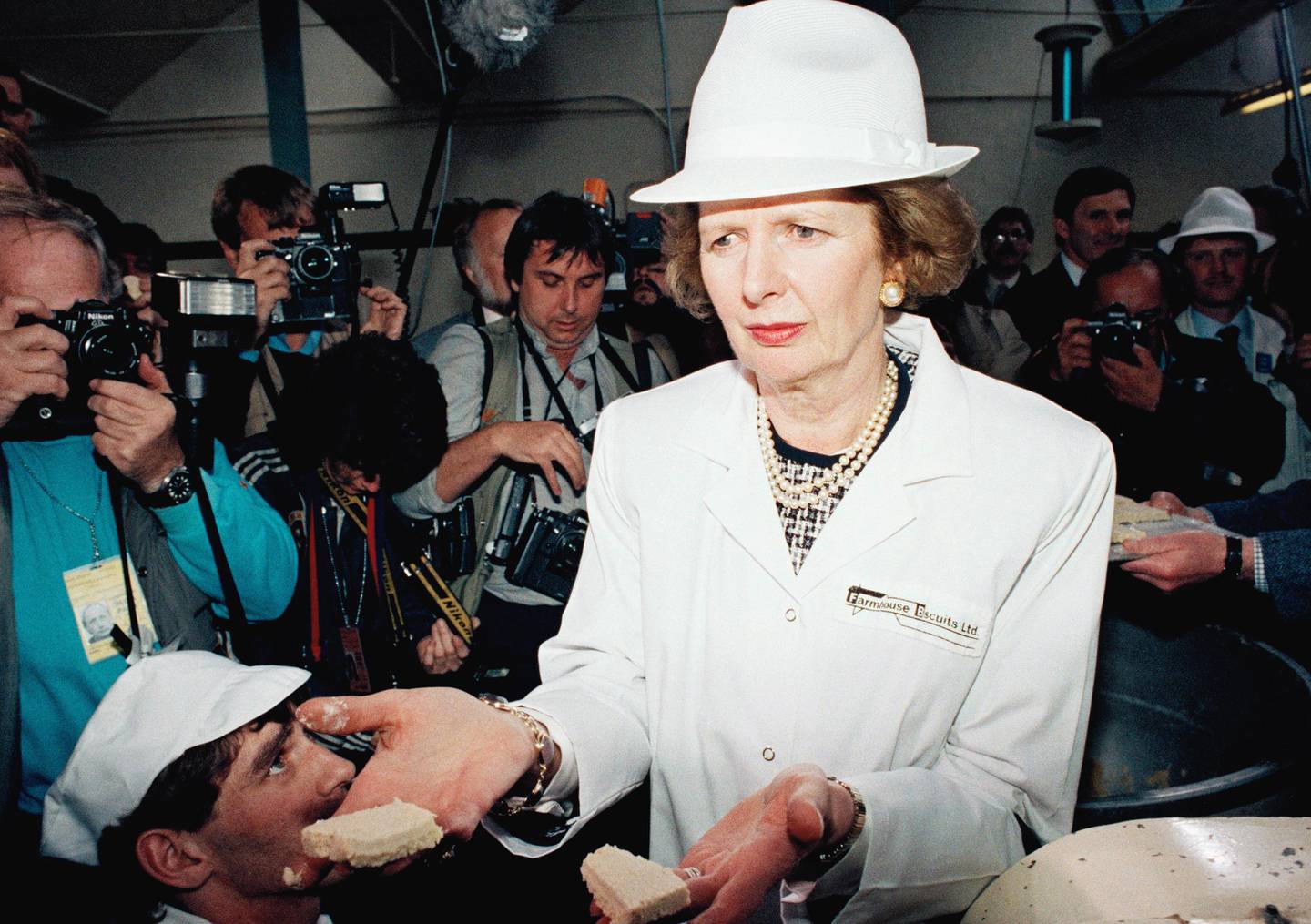 Britain?s Prime Minister Margaret Thatcher, wearing a white trilby hat and coat, hands out biscuits to photographers while touring the Farmhouse Biscuit factory at Nelson, Lancashire on Friday, May 23, 1987, during a stop on her election campaign tour. Britain goes to the polls in a General Election on June 11th. (AP Photo/Dave Caulkin)