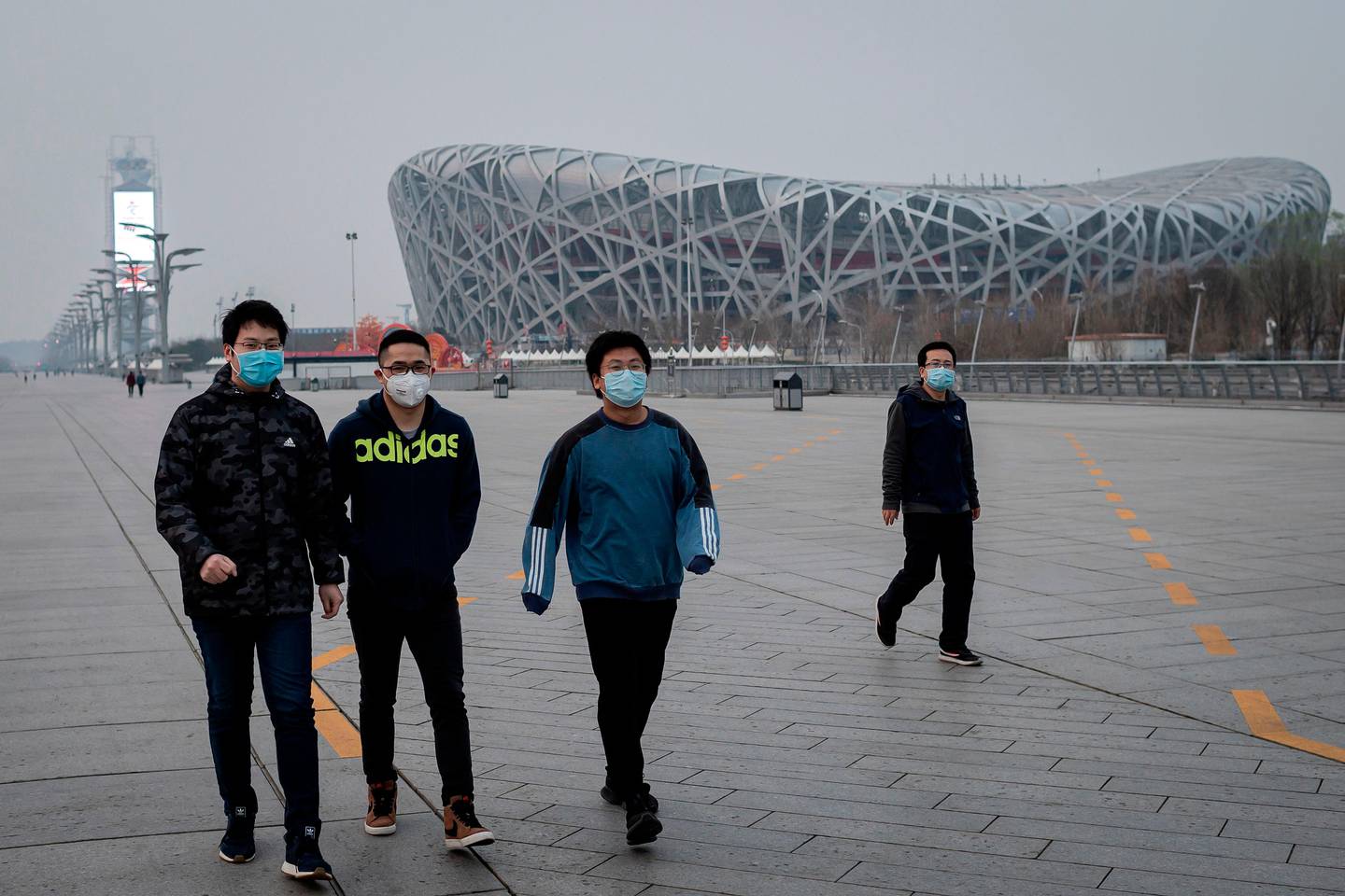 People wearing facemasks amid concerns of the COVID-19 coronavirus walk at the Olympic park outside the national 'Birds Nest' stadium (R), the site of the 2008 Beijing Olympics, in Beijing on March 24, 2020. - The 2020 Tokyo Olympics have been postponed to no later than the summer of 2021 because of the coronavirus pandemic sweeping the globe, the International Olympic Committee announced on March 24. (Photo by NICOLAS ASFOURI / AFP)