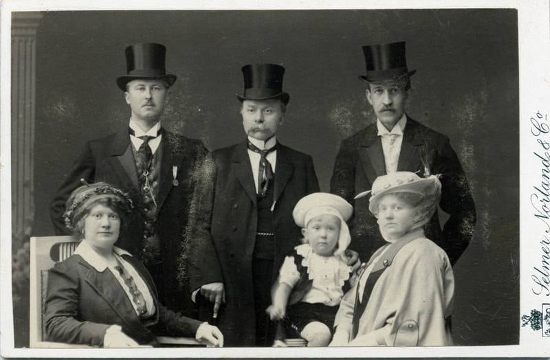 Mannen i midten med flosshatt er snekker Nils Johansson. Hans datter Olga Kamilla Josefine foran til høyre, hennes mann, Hartvig Fosnes Hansen bak henne. På fanget har hun sin førstefødte sønn Harry. De andre mennene på bildet er ikke kjent. Bildet er tatt i 1914.