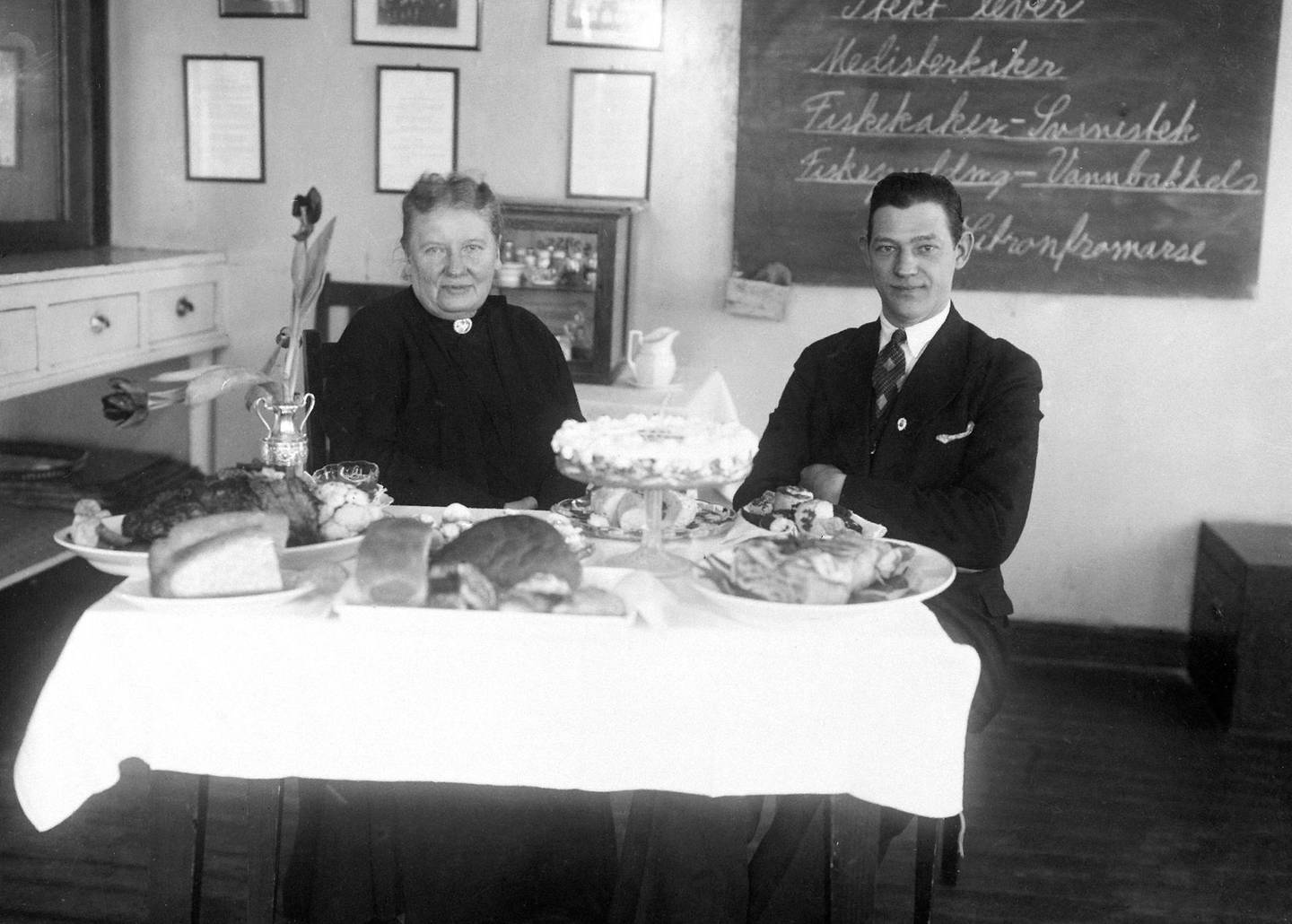 Henriette Schønberg Erken med bestyrer John Hannevig Olsen. Schønberg Erken viste frem kokkekunsten, med beskrivelser bak på tavlen som «Fiskekaker - Svinestek» og «Fiskepudding - Vannbakkels». FOTO: ANNO DOMKIRKEODDEN