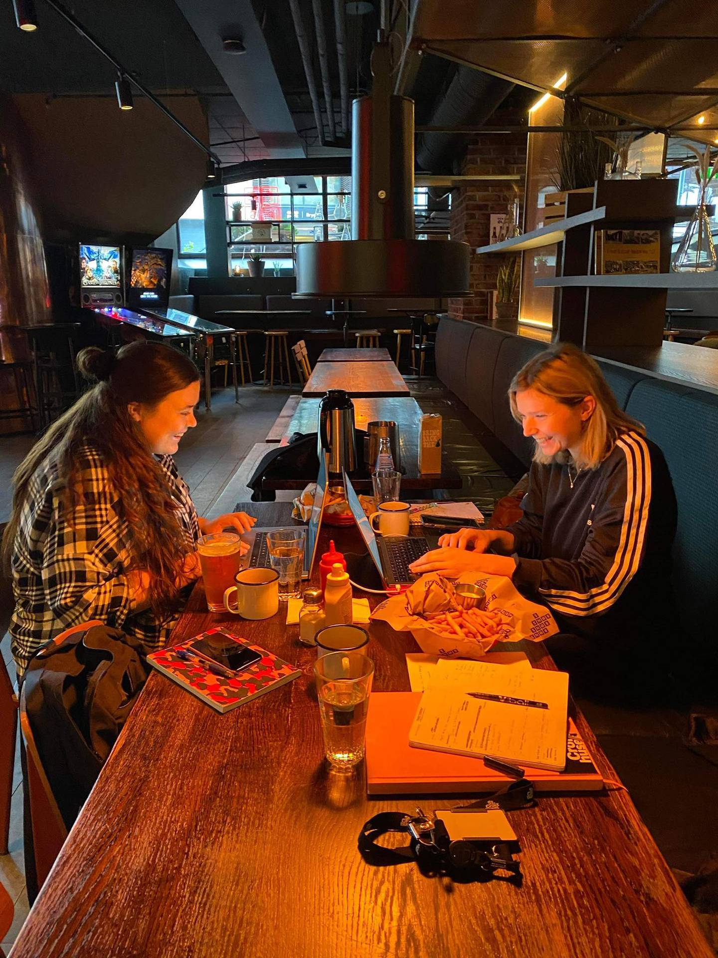 To kvinner sitter ved et bord fullt av papirer, på baren Tæps. De ser inn i hver sin pc og smiler.