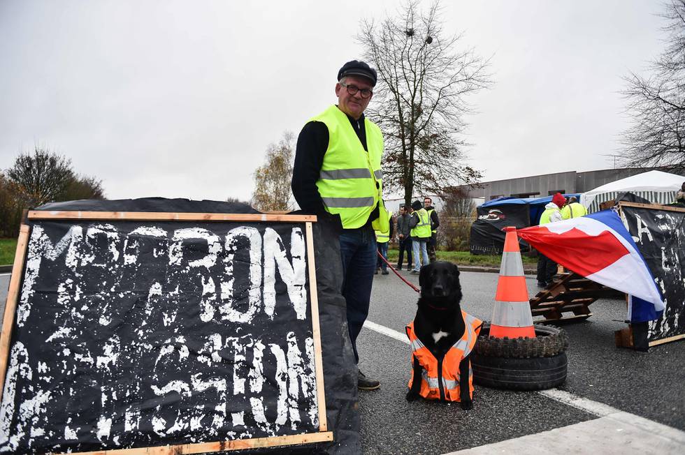 A Yellow Vest (Gilets jaunes) protester poses with his dog in front of a sign that reads in French, 'Macron (referring to the French president) Resignation', blocking part of the access to the Martin Bower warehouse which is responsible for the deliveries of the American fast food franchise McDonald for the Western France region on December 2, 2018, at Torce near Rennes, western France. - The "yellow vest" or "Gilets jaunes" movement is named after the high-visibility jackets which motorists are required to carry in their cars. The movement, organised through social media, has steadfastly refused to align with any political party or trade union and includes many pensioners and has been most active in small urban and rural areas where it has blocked roads, closed motorway toll booths, and even walled up the entrance to tax offices. (Photo by JEAN-FRANCOIS MONIER / AFP)