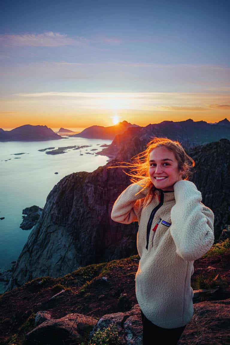 Jente i 20-årene med brunt hår, iført en hvit ullgenser. Hun står på et fjell med vann og en solnedgang i bakgrunnen.