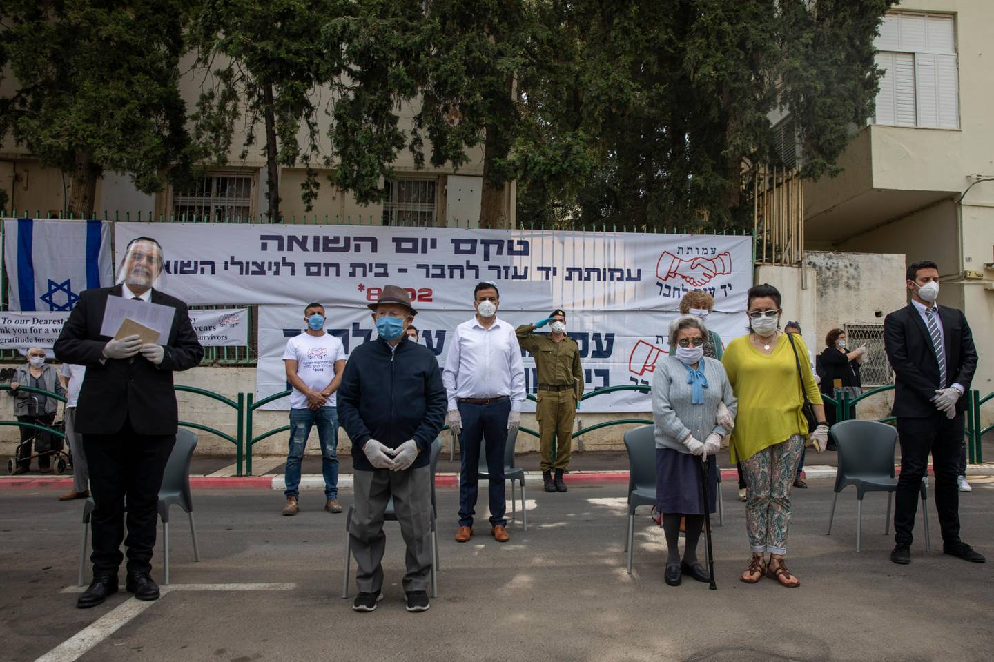 Holocaust survivor Shalom Stamberg, second left, and Holocaust survivor Miriam Linial, third right, wear a mask and keep a safe distance from others while the two-minute-long siren sounds during an annual Holocaust memorial ceremony held outside this year because of the coronavirus, in the northern Israeli city of Haifa, Israel, Tuesday, April 21, 2020. Stamberg, 97 and Linial, 99, both Holocaust survivors from Poland. The Hebrew sign reads "Holocaust memorial ceremony and Yad Ezer La-Haver foundation, warm home for Holocaust survivors". (AP Photo/Ariel Schalit)
