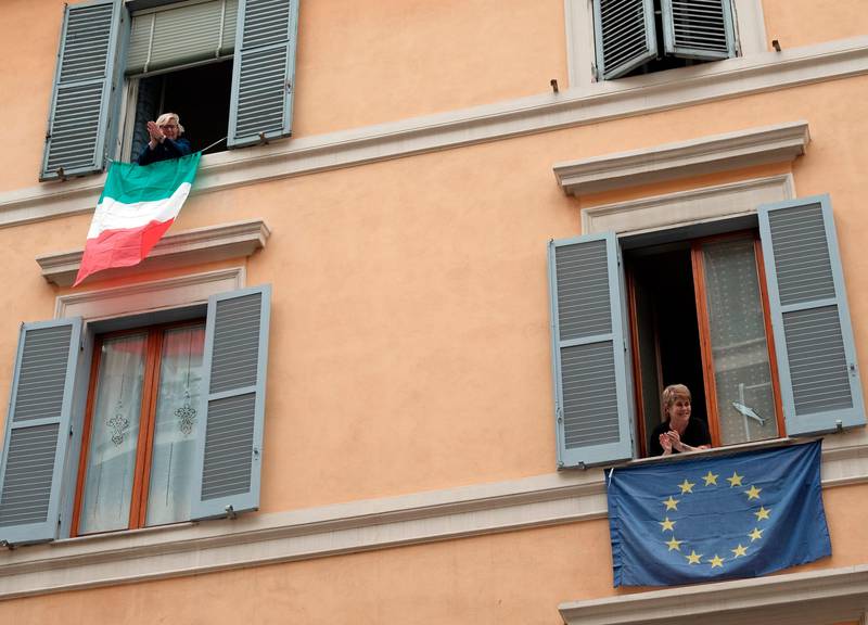 People clap their hands as the Italian, top, and European Union flags hang from windows, in Rome, Saturday, March 14, 2020. At noon in Italy, people came out on their balconies, terraces, gardens or simply leaned out from open windows to clap for several minute in a gesture of thanks for the doctors, nurses, other hospital staff and ambulance crews who have been treating and helping infected patients. (Mauro Scrobogna/LaPresse via AP)
