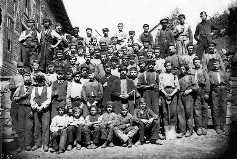 Arbeidere ved Kværner Brug cirka 1870, for anledninga med hodeplagg. Det finnes også et bilde der alle er barhoda. Legg merke til alderen på gutta i første rekke.