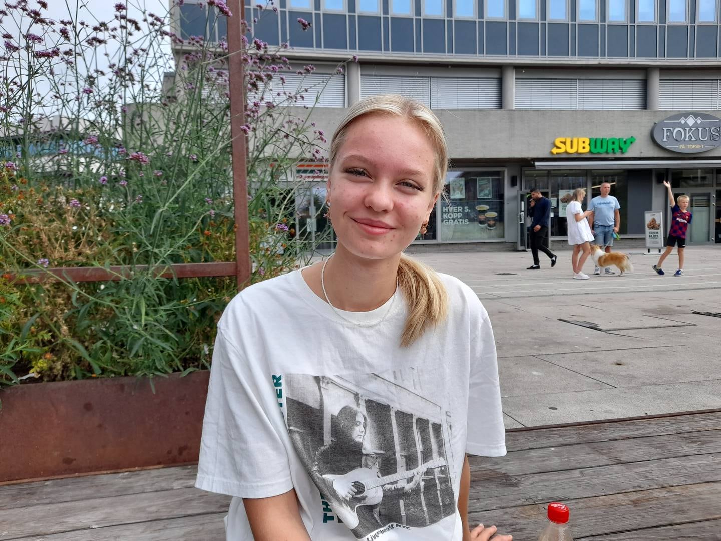 En ung kvinne i hvit t-skjorte, smiler, på benk på Stortorvet, Fredrikstad