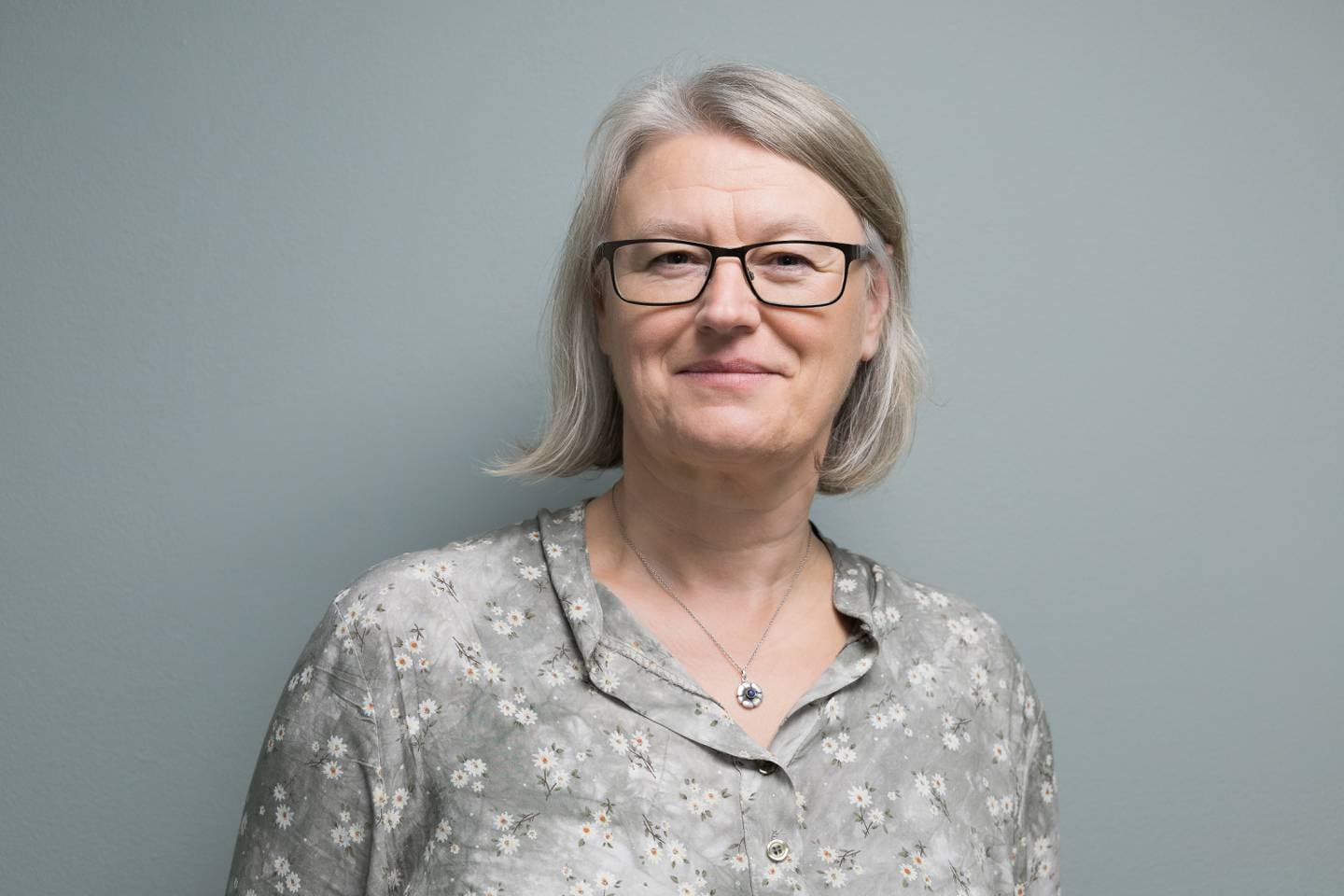 Pressebilde av Kjersti Margrethe Rydland i en småblomstrete grå bluse foran en lys vegg.