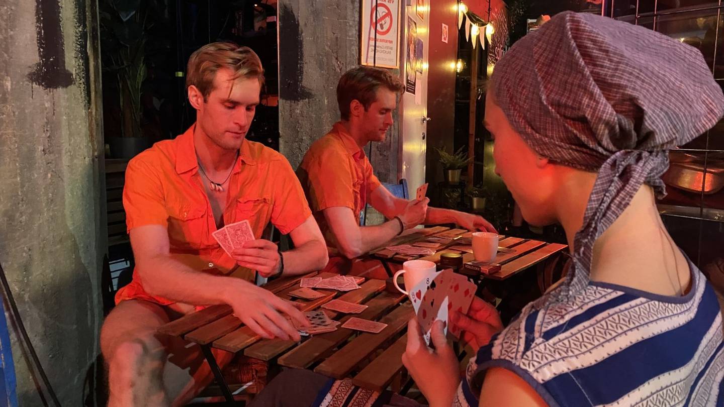 Mann i 20-årene iført oransje skjorte spiller kort med kvinne i 20-årene iført skaut og stripete skjorte. De sitter ved et brunt bord på settet til Mamma Mia.