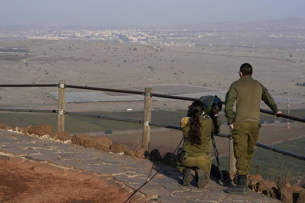 To personer - en sittende og en stående i militærklær med ryggen til, ser innover et åpent landskap med hus og bebyggelse i det fjerne.