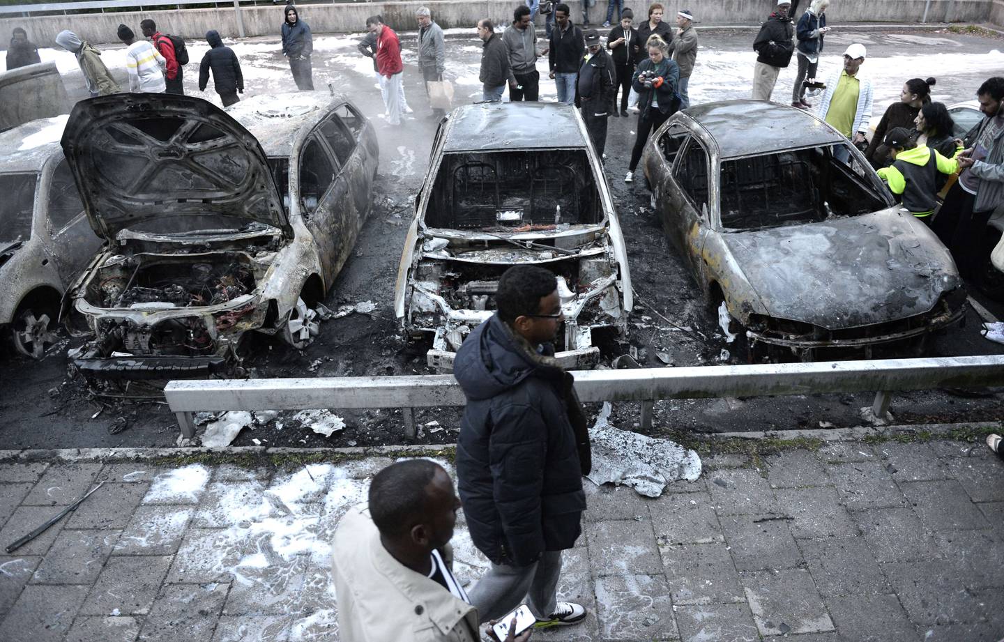 People inspect the scene where cars stand gutted by fire in the Stockholm suburb of Rinkeby after youths rioted in several different suburbs around Stockholm, Sweden for a fourth consecutive night on May 23, 2013. In the suburb of Husby, where the riots began on Sunday in response to the fatal police shooting of a 69-year-old machete-wielding man, 80 percent of residents are immigrants and the unrest has highlighted Sweden's failure to integrate swathes of its immigrant population, but in this small, consensus-driven country, there was little agreement on how to solve the problem.  AFP PHOTO / JONATHAN NACKSTRAND