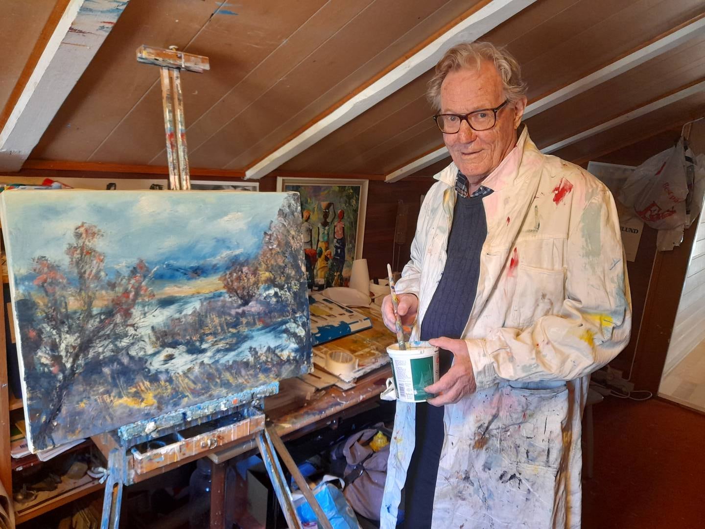 Øyvind Nordby, i malerfrakken sin, holder en pensel i hånden. Foran han står et maleri på et staffeli.